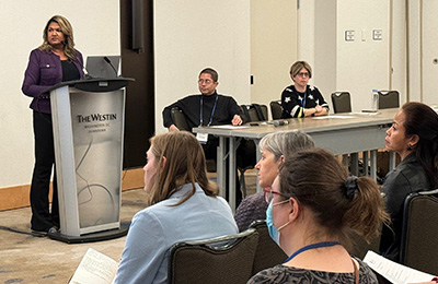 Image of a woman presenting at a podium with two people sitting alongside her in a panel discussion with the audience looking on.